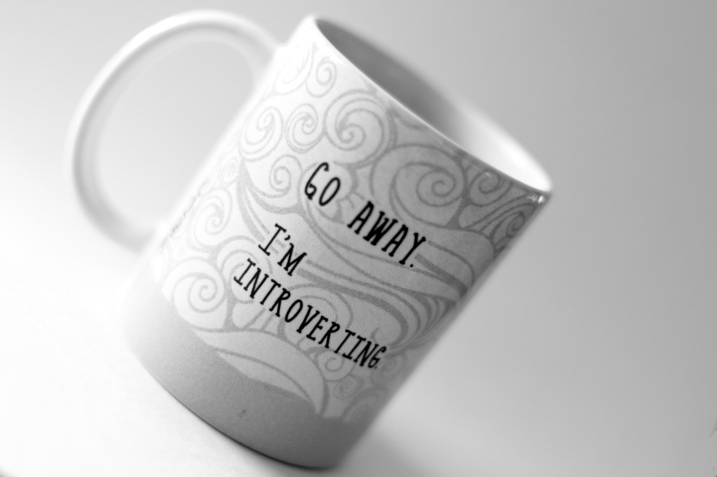 Black and white image of a tilted mug that says "GO AWAY. I'M INTROVERTING." in block letters against an abstract swirly design.